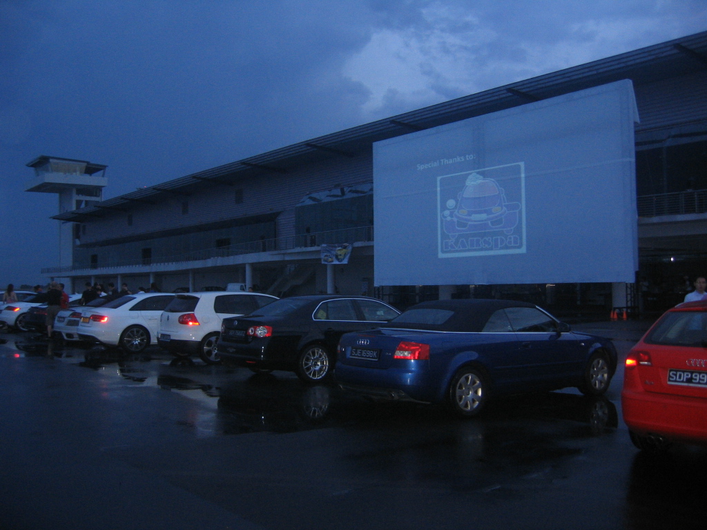 Drive-in event at Changi Exhibition Centre with cars and large screen under moody sky.
