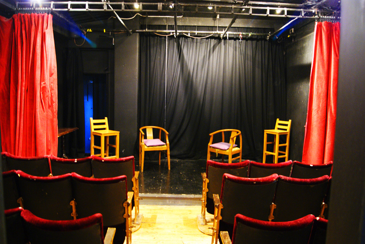 Intimate theater setup with wooden chairs and red curtains for workshops and discussions.