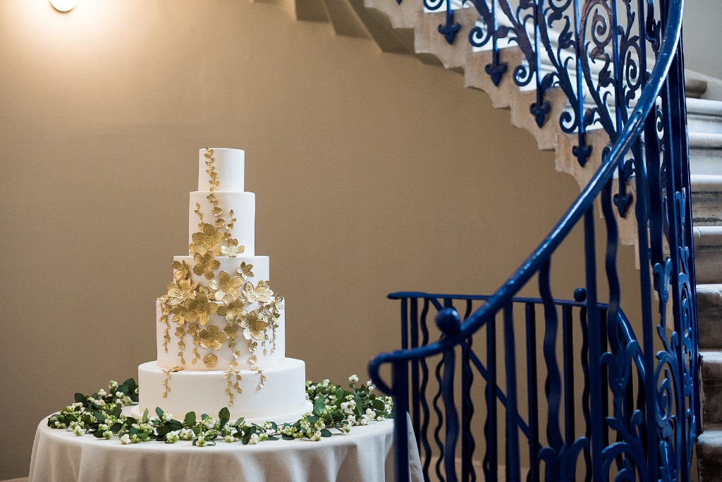 Elegant wedding cake in The Great Hall, Queen's House, with ornate staircase backdrop.
