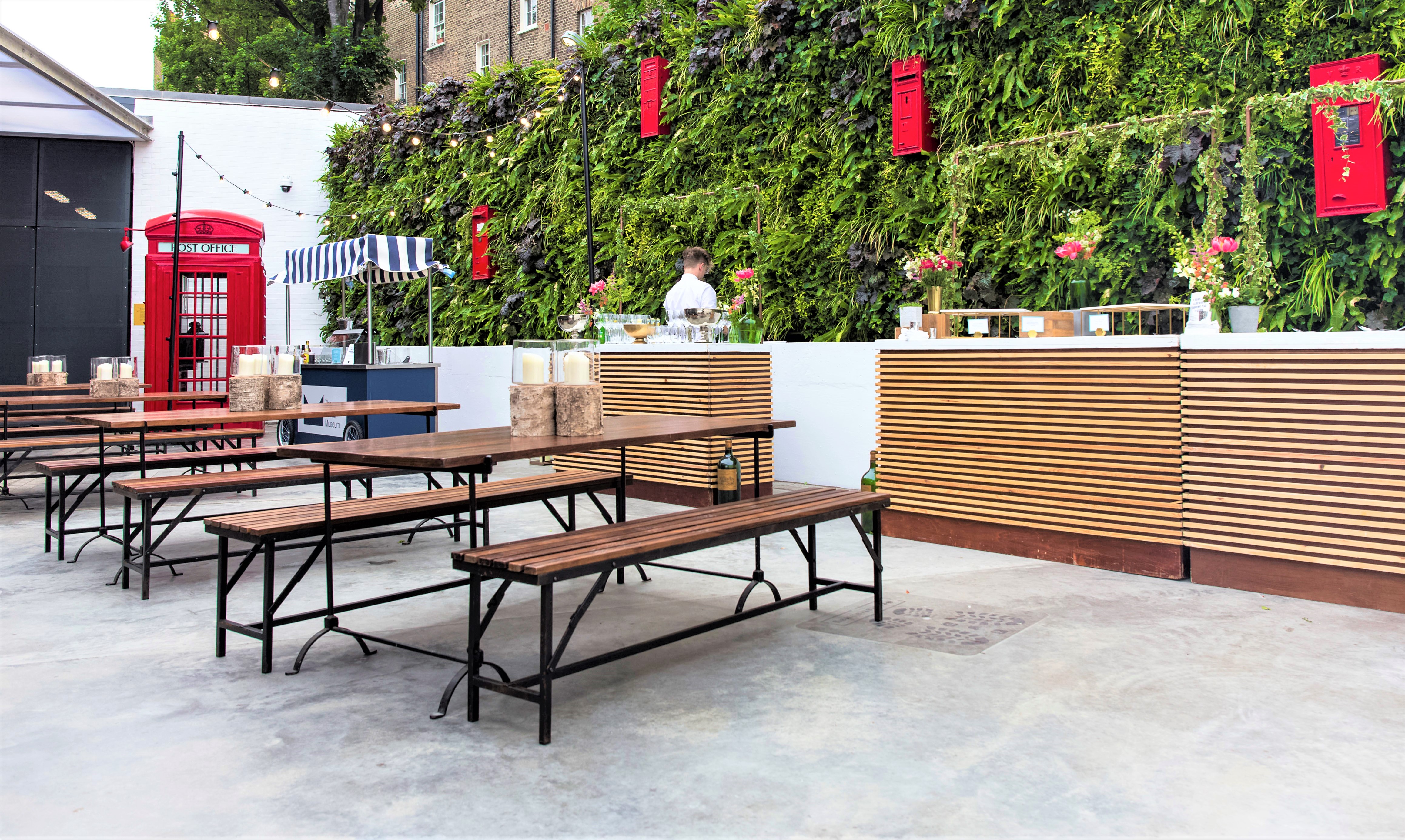 Vibrant outdoor event space at The Postal Museum with rustic benches and lush greenery.
