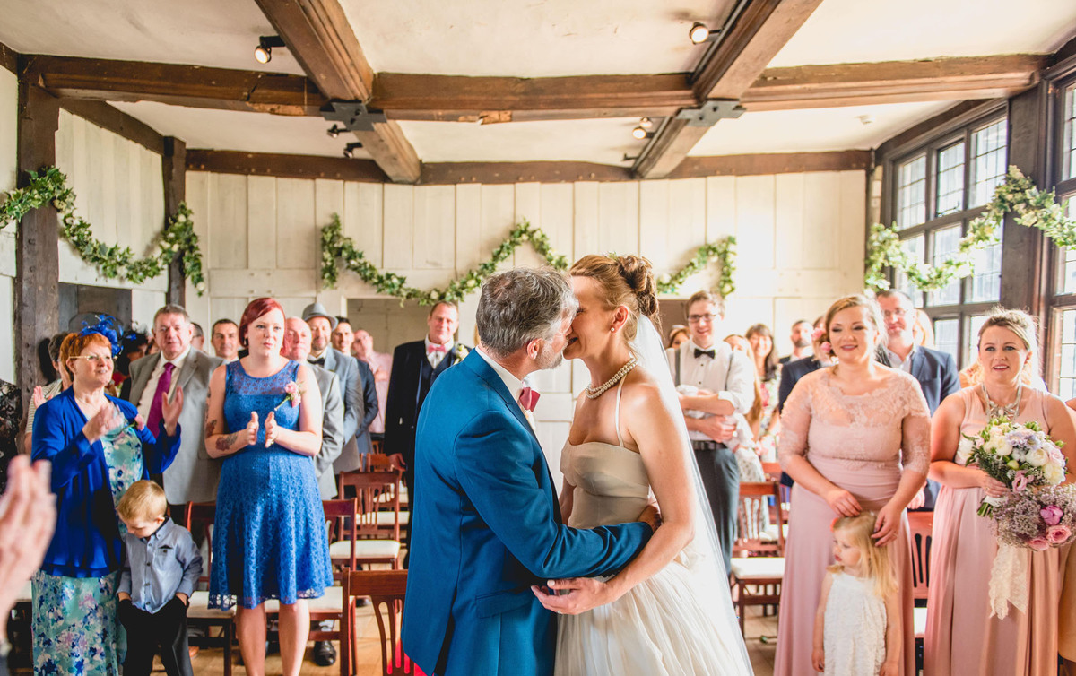 Wedding ceremony in Blakesley Hall's Great Hall, featuring greenery and couple's first kiss.