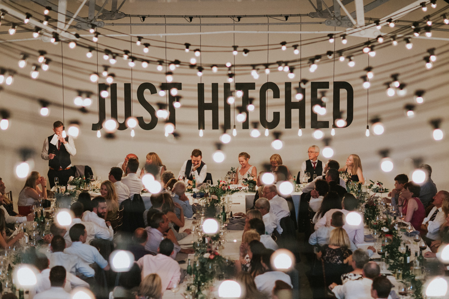 Wedding reception in Shoreditch Studios with "JUST HITCHED" backdrop and string lights.