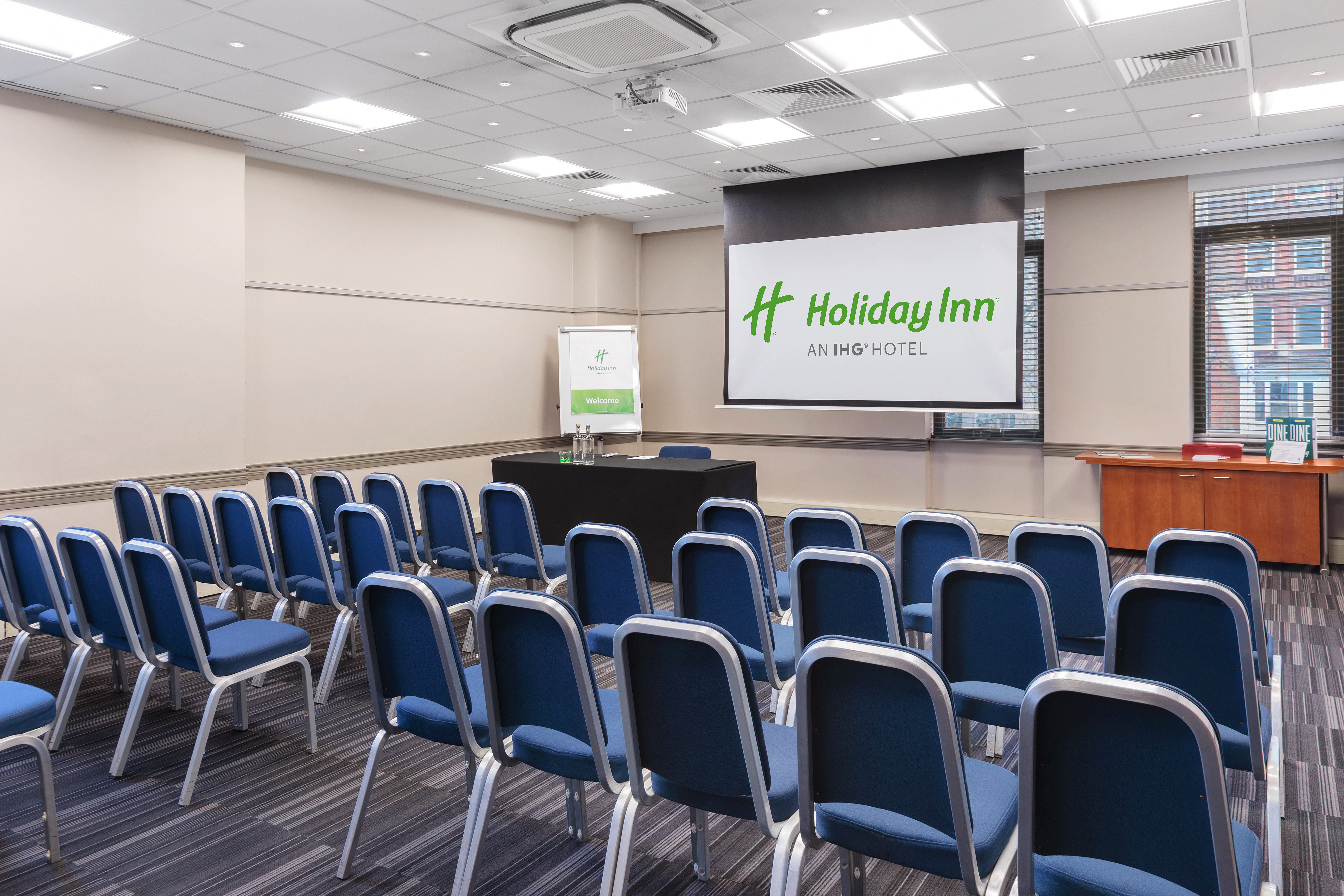 Meeting room at Holiday Inn London Kensington with blue chairs and projection screen.
