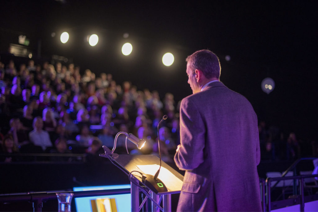 Speaker addressing audience in Millennium Point auditorium during a professional conference.