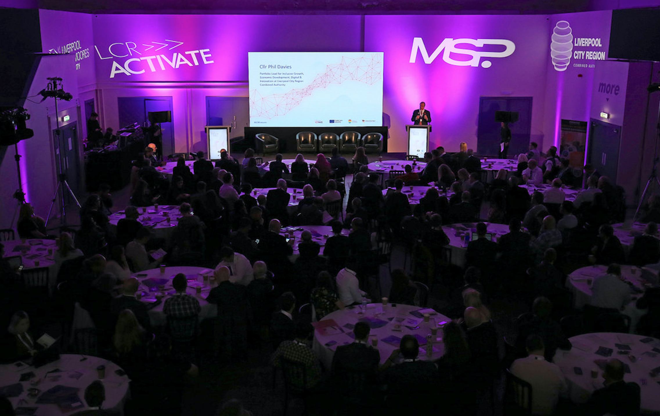 "Event Hall in Hinterlands with speaker at podium, purple lighting for a conference."