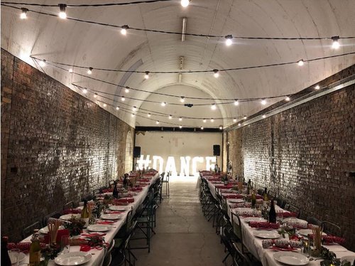 Long dining table in industrial venue for dining and dance events, illuminated by string lights.