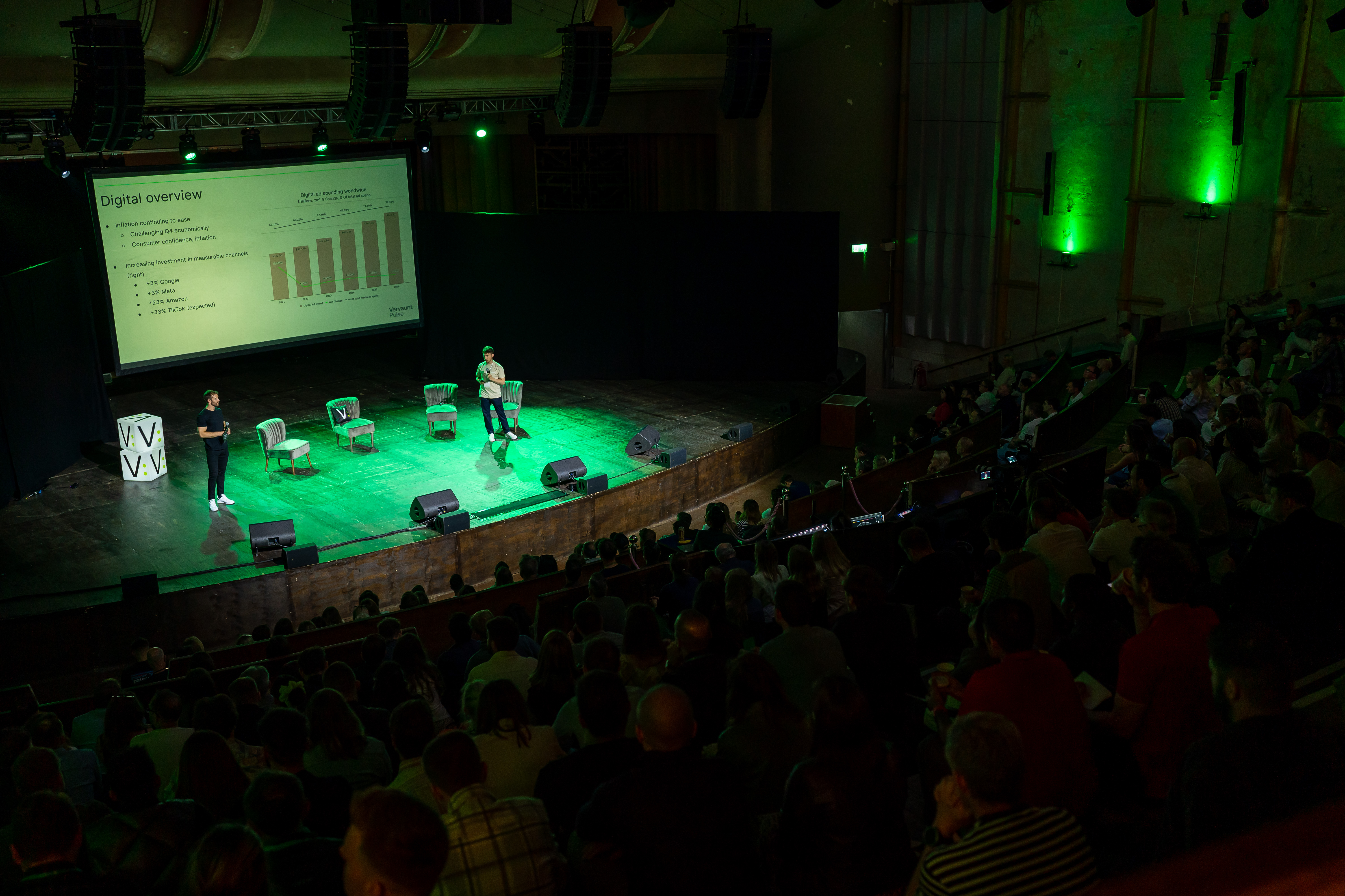 Conference speaker engaging audience in EartH Theatre with green lighting and data display.