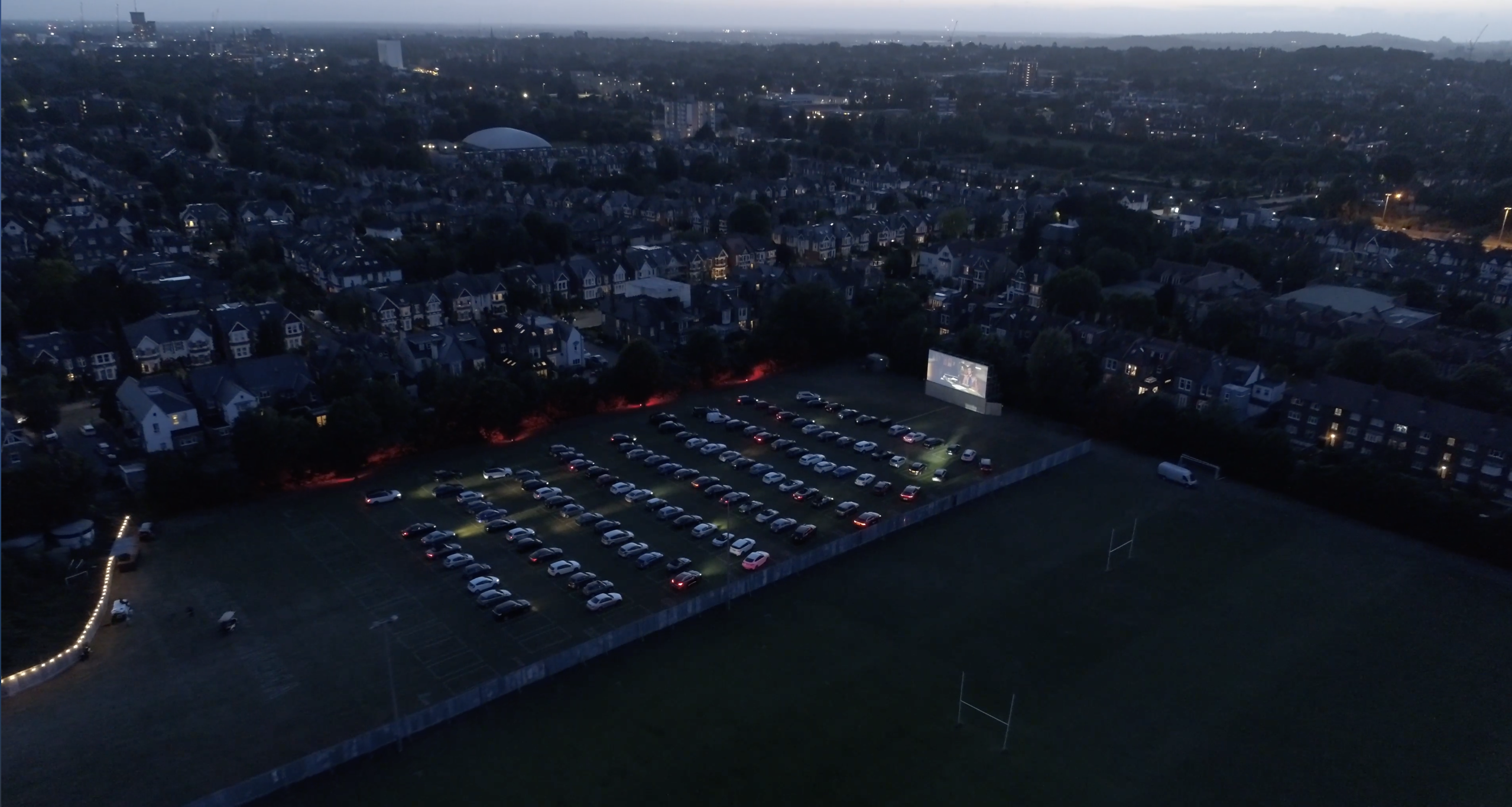 Outdoor drive-in movie setup at Sunset Cinema with parked cars and large screen.