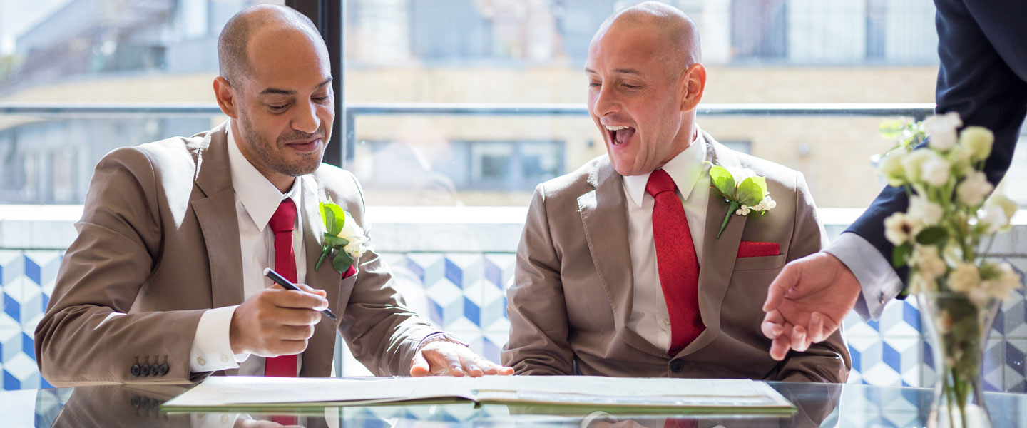 Two grooms in stylish suits joyfully signing wedding documents at Wallacespace Clerkenwell.