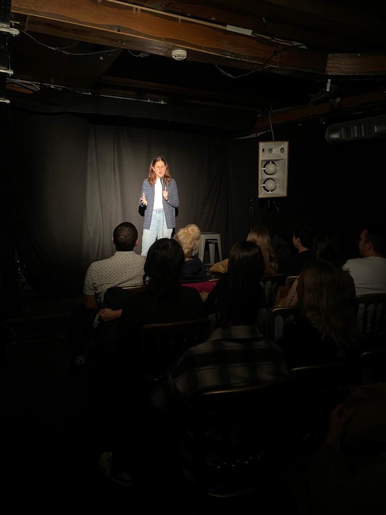 "Performer on stage in cozy Hoxton Cabin Basement for stand-up comedy event."