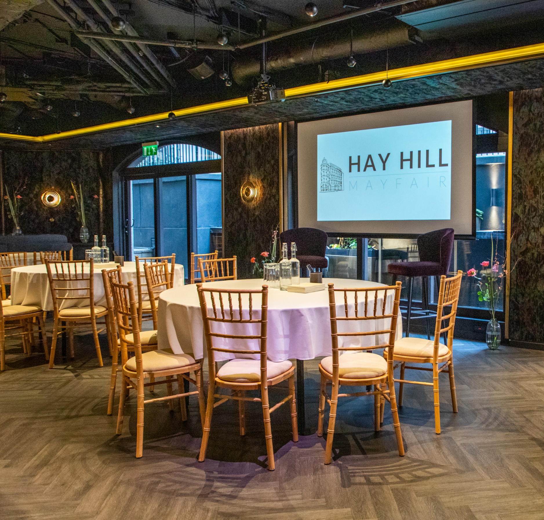 Elegant event space at The Bar in Mayfair with round tables and branded screen. 