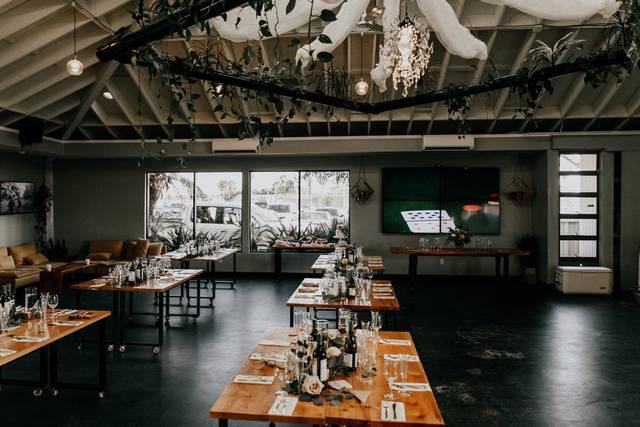 Indoor event space with long wooden tables, greenery, and natural light for gatherings.