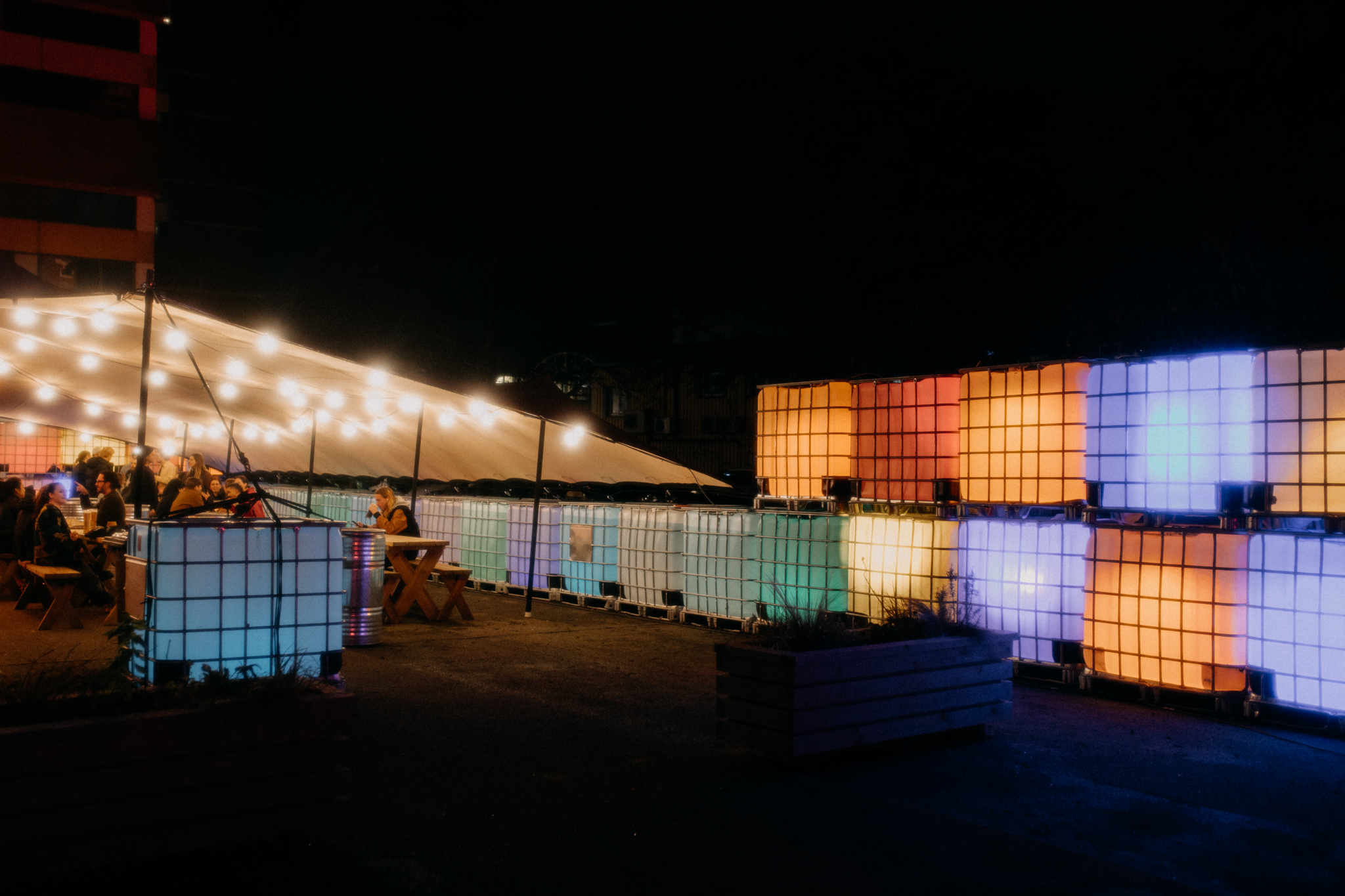 Outdoor event space with illuminated containers and string lights for gatherings.