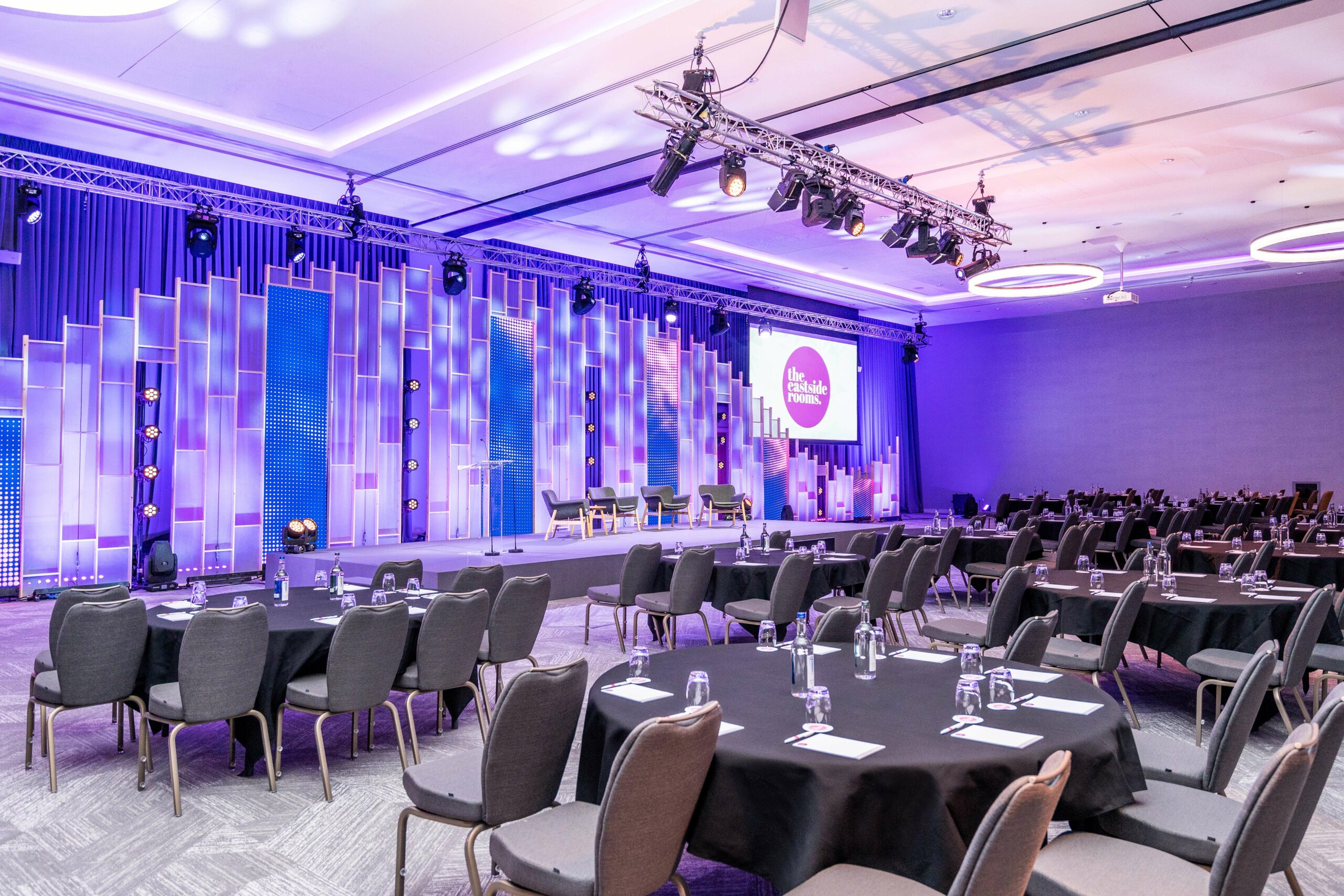 Conference space with round tables and purple-lit stage for networking events.