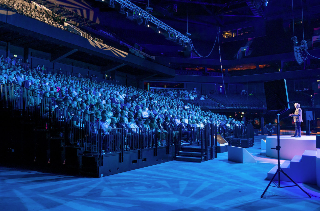 Speaker on stage at Co-op Live arena during an engaging seminar with vibrant blue lighting.