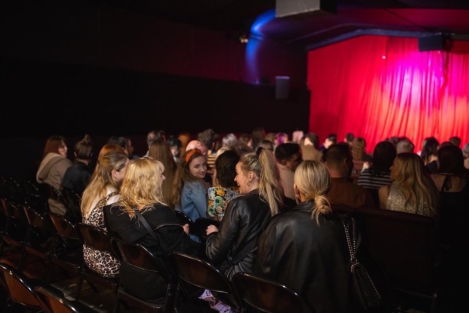 Engaged audience in GRUB MCR venue with vibrant red backdrop for community events.