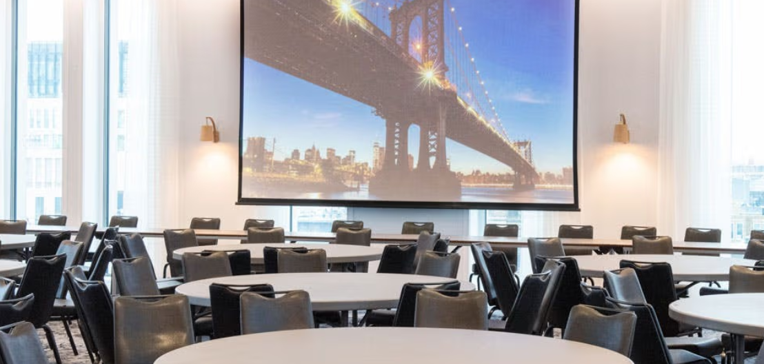 Versatile meeting space in voco Manchester with round tables and projection screen.