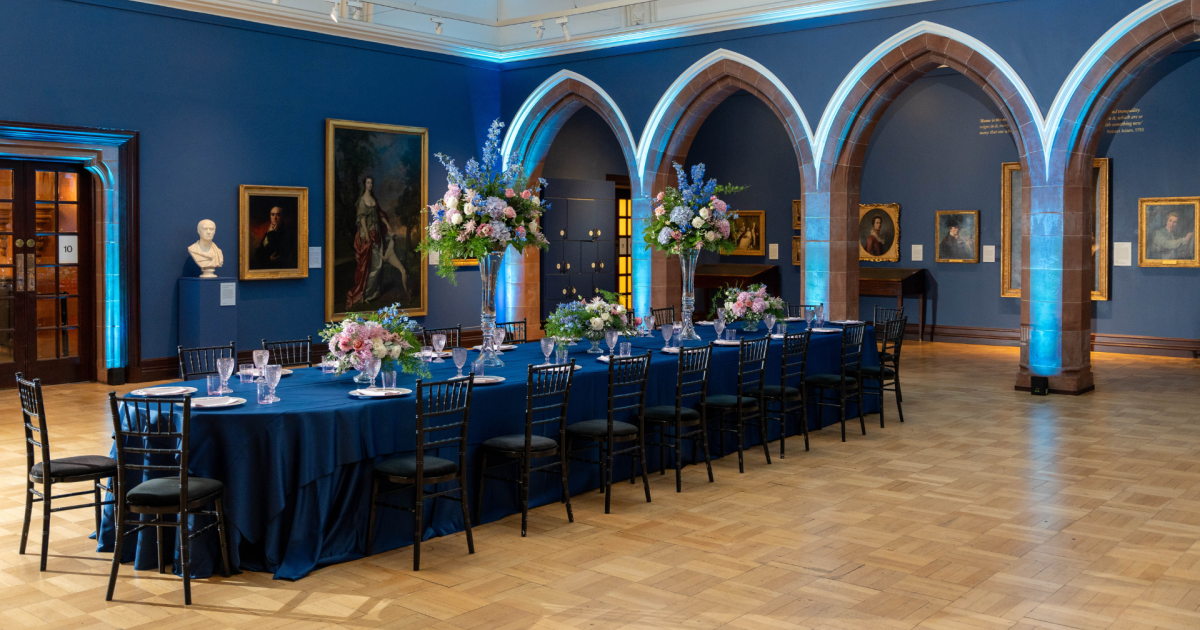 Catered gallery banquet with blue uplighting and floral centerpieces for corporate event catering.