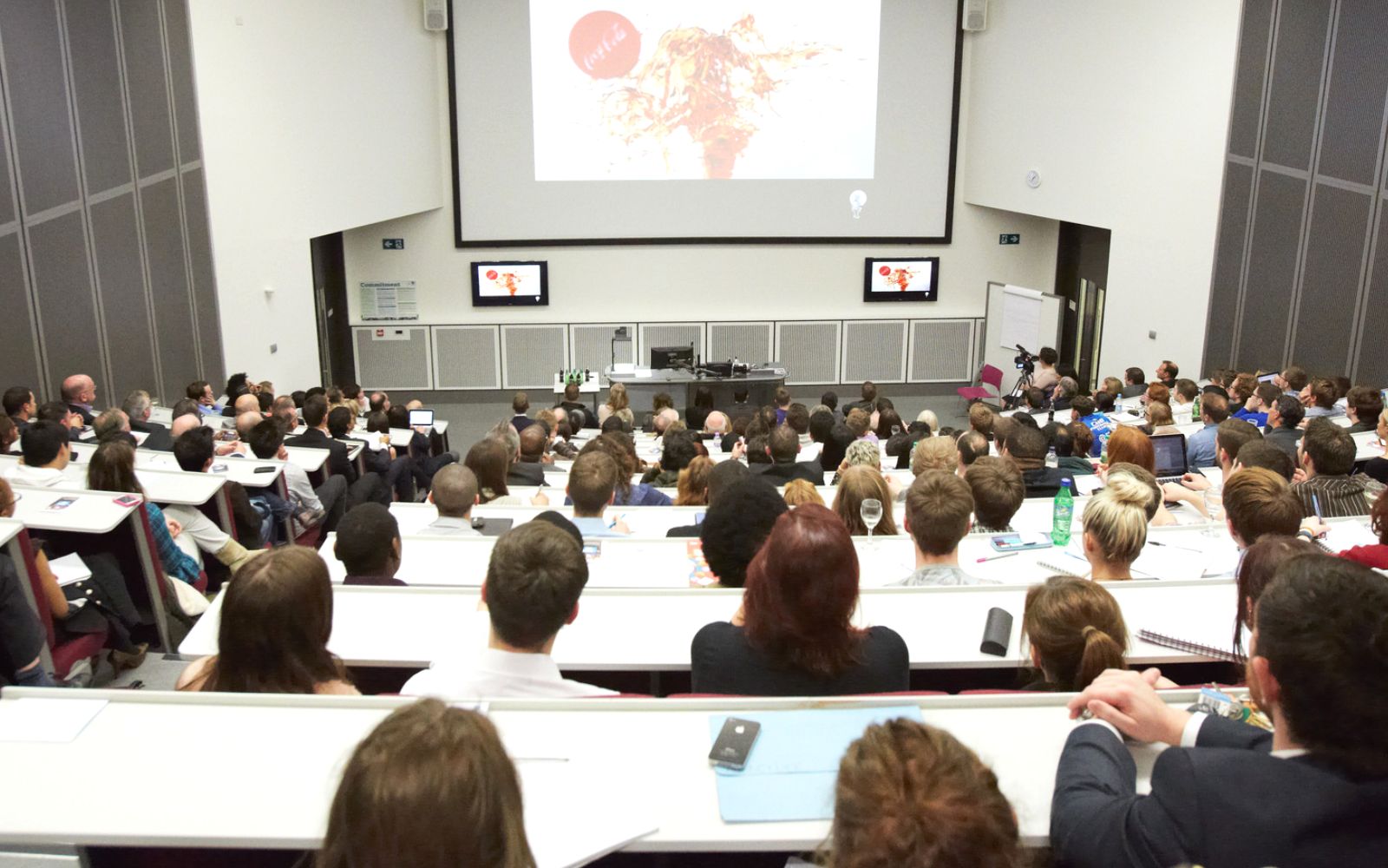 Engaged audience in modern business school seminar with tiered seating and tech displays.