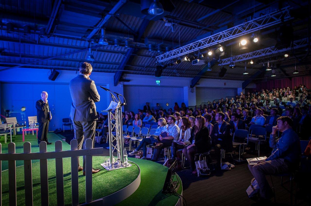 "Conference speaker at podium in green-themed Business Design Centre auditorium"